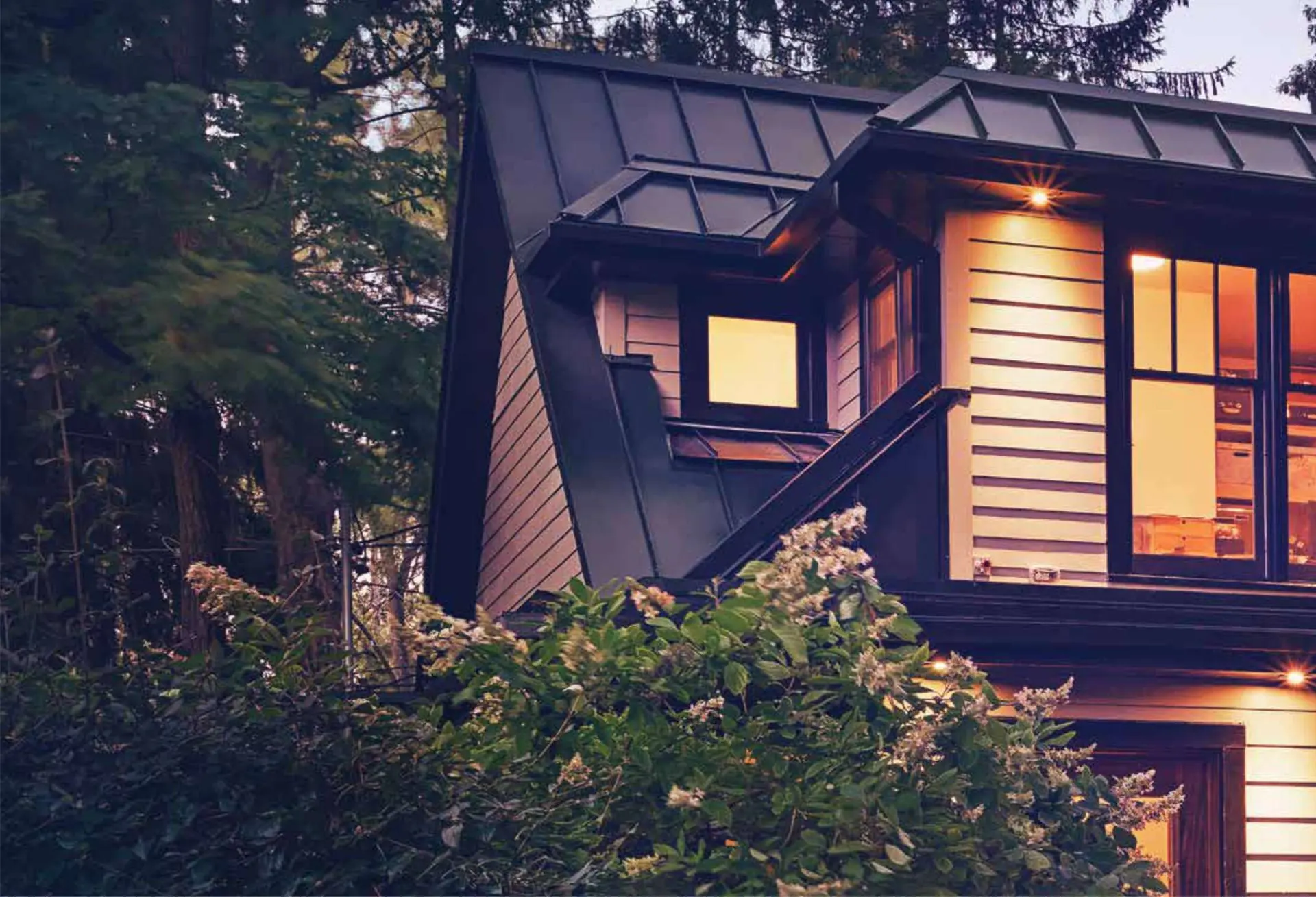 A dark standing-seam metal home glowing warmly at dusk among trees